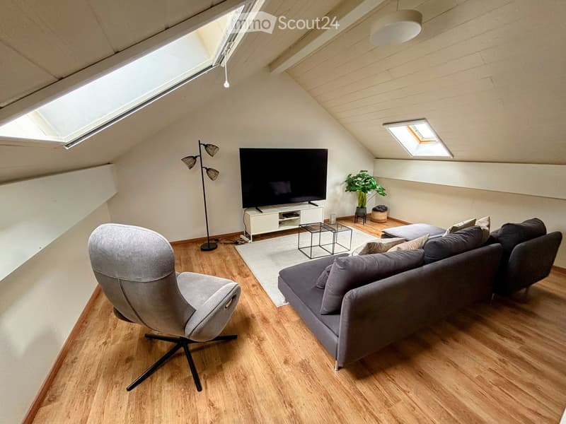 Modern living room with wooden flooring, grey sofa, armchair, television, small table, floor lamp, and a potted plant.