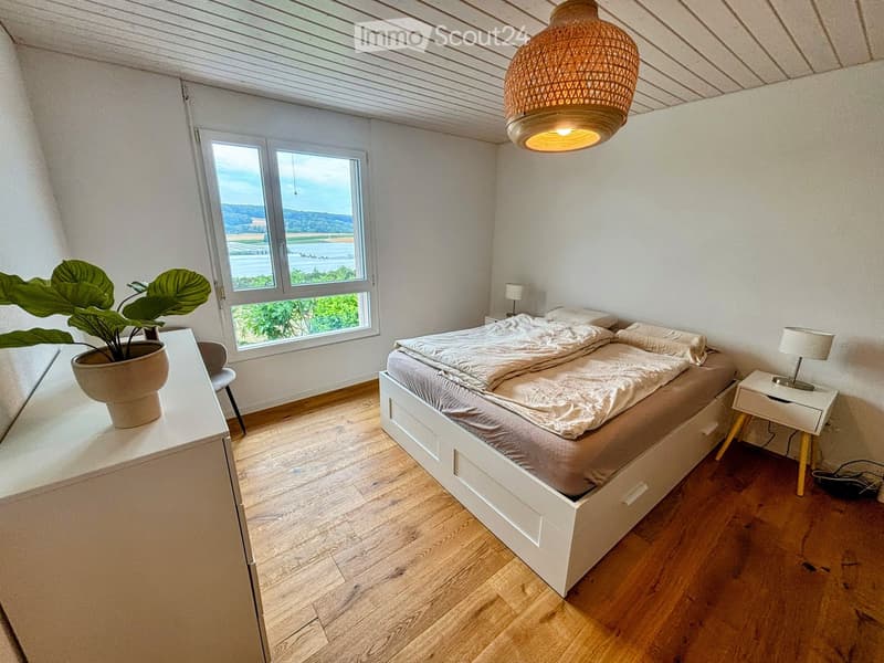 Bright bedroom with wooden flooring, large window offering scenic view, white bed with grey bedding, nightstand with lamp, wicker pendant light, and potted plant on side table.