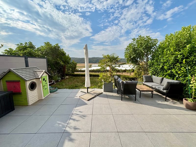 A well-maintained garden with a tiled patio, outdoor furniture including a sofa, chairs, and a table, a large umbrella, and a decorative dog house.