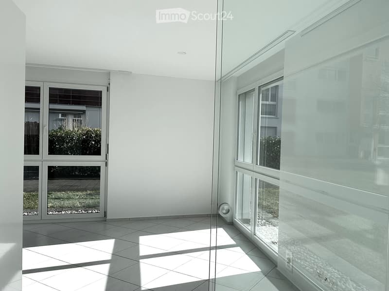 A modern empty room with white walls and tiled floor, large windows allowing natural light, and glass walls providing transparency.