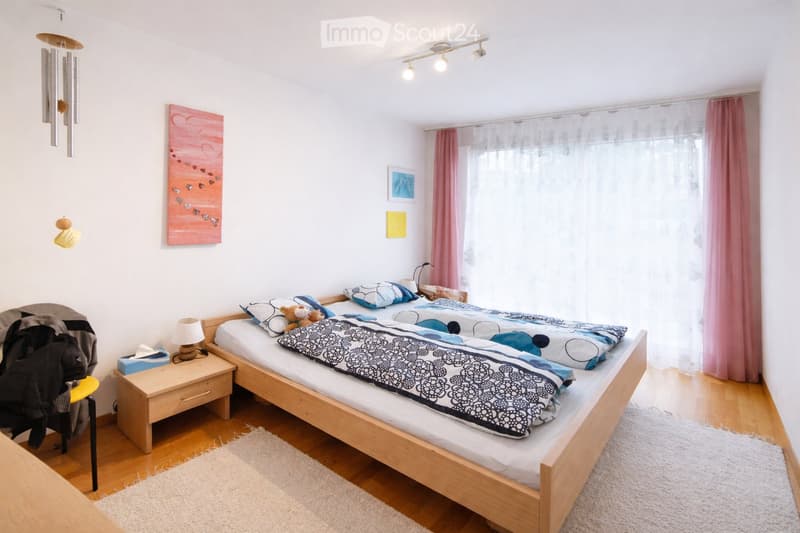 Cozy bedroom with wooden bed, nightstand, lamp, and decorative items. Features a large window with pink curtains and artwork on the walls.