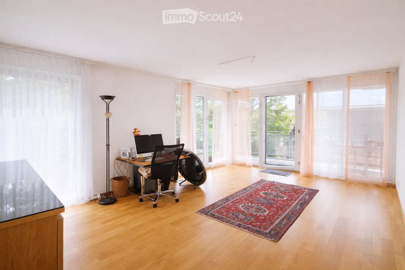 Brightly lit room with wooden flooring, large windows with sheer curtains, a desk with a computer and chair, a floor lamp, and a patterned rug.