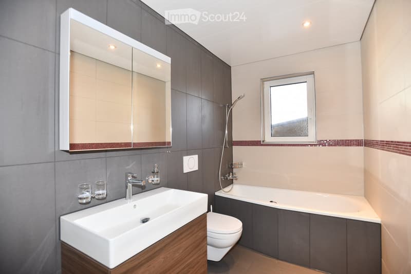 Modern bathroom with a white sink, wooden cabinet, large mirror, bathtub, toilet, and showerhead. The walls are tiled in gray and beige with a red accent border. A small window allows natural light.
