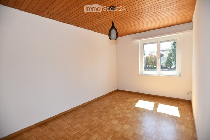 Empty room with wooden flooring, white walls, and a wooden ceiling. A single window with white frames allows natural light to enter, and a modern pendant light hangs from the ceiling.