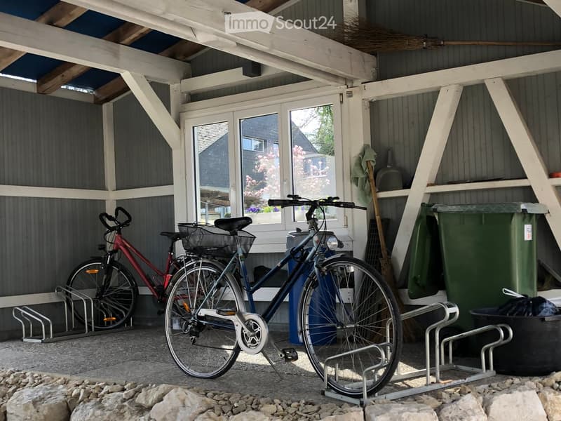 A covered outdoor area with two bicycles parked on racks, a broom, a green trash bin, and a view of a house with windows and a garden in the background.
