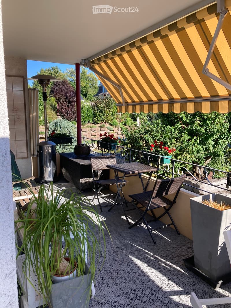 Balcony with a yellow striped awning, a small table with two chairs, a wicker bench, a patio heater, and potted plants.