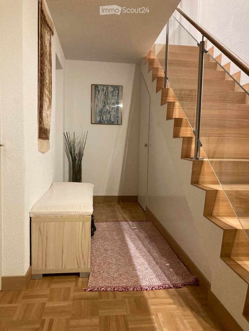 Modern hallway with wooden flooring, a staircase with glass railing, a small bench, a vase with dried plants, and a framed artwork on the wall.