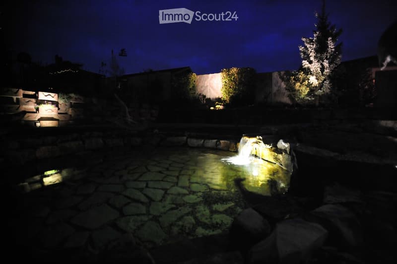 Nighttime view of a garden with a small illuminated water feature and surrounding trees and shrubs.