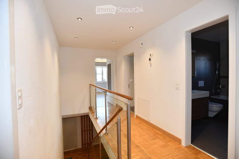 Modern hallway with wooden flooring, glass railing staircase, recessed ceiling lights, and a view into a bathroom with dark tiles and fixtures.
