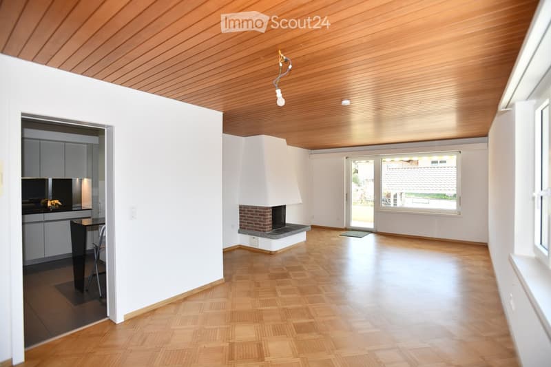 Bright living room with wooden flooring, white walls, and a central brick fireplace. A small kitchen area is visible with cabinets and a countertop. A single chair and table are placed near the kitchen.