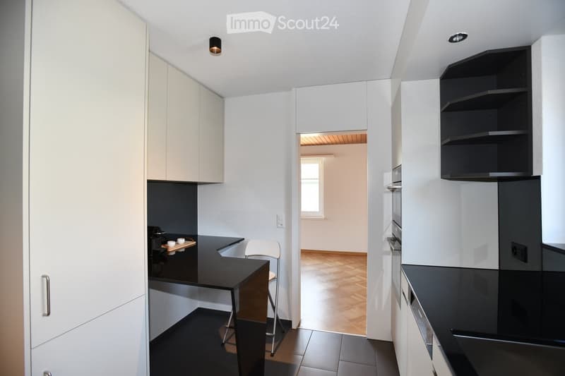 Modern kitchen with white cabinets, black countertops, and built-in appliances. A small table with a stool is visible, and a doorway leads to another room with wooden flooring.