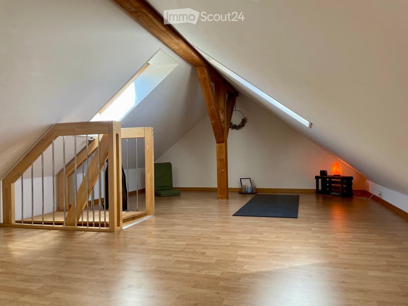 Bright attic room with wooden floors, slanted ceilings, and skylights. Features a small table with a lamp, a cushioned seat, and a staircase leading to another level.