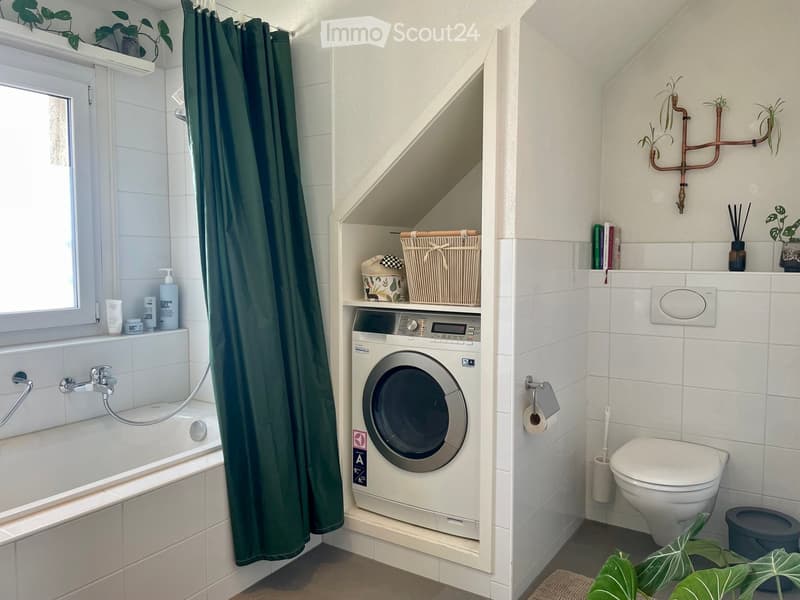 Modern bathroom with a bathtub, shower curtain, washing machine, toilet, and decorative plants. Includes a window with a shelf for toiletries and a small storage area above the washing machine.
