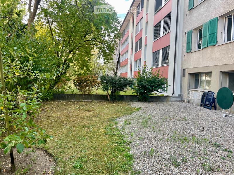Garden area with gravel pathway, small grassy patch, outdoor furniture including chairs and a round table, surrounded by trees and buildings.