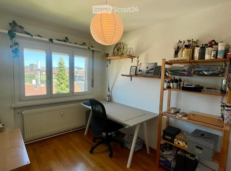 Well-lit study or office space with a modern desk, ergonomic chair, wooden shelving unit, and a large window offering a city view.
