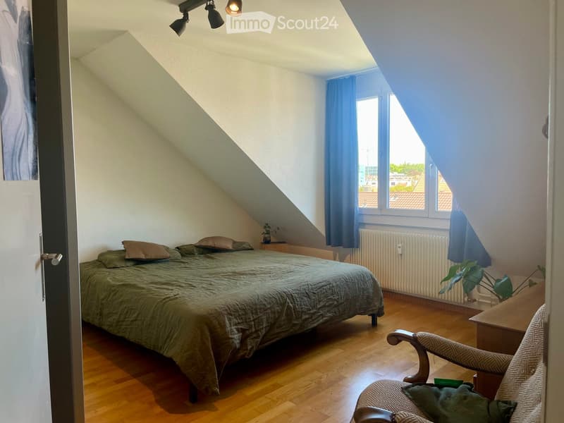 Cozy attic bedroom with wooden flooring, slanted ceilings, and large windows. Features a bed with green bedding, a wooden chair, a small table, and a plant near the window.