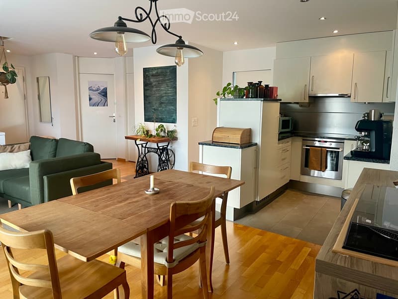Open-plan living and dining area with a green sofa, wooden dining table, and modern kitchen featuring stainless steel appliances and white cabinets.