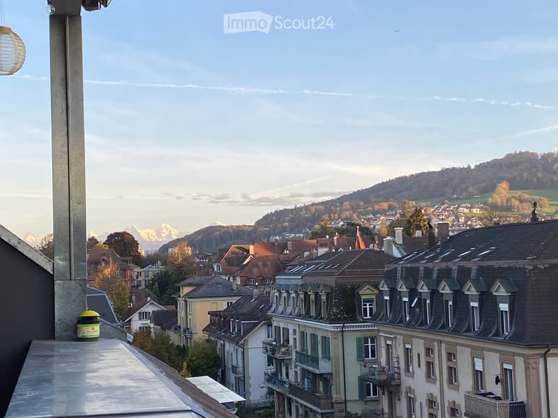 View from a balcony showing a cityscape with traditional houses, green hills, and distant mountains.