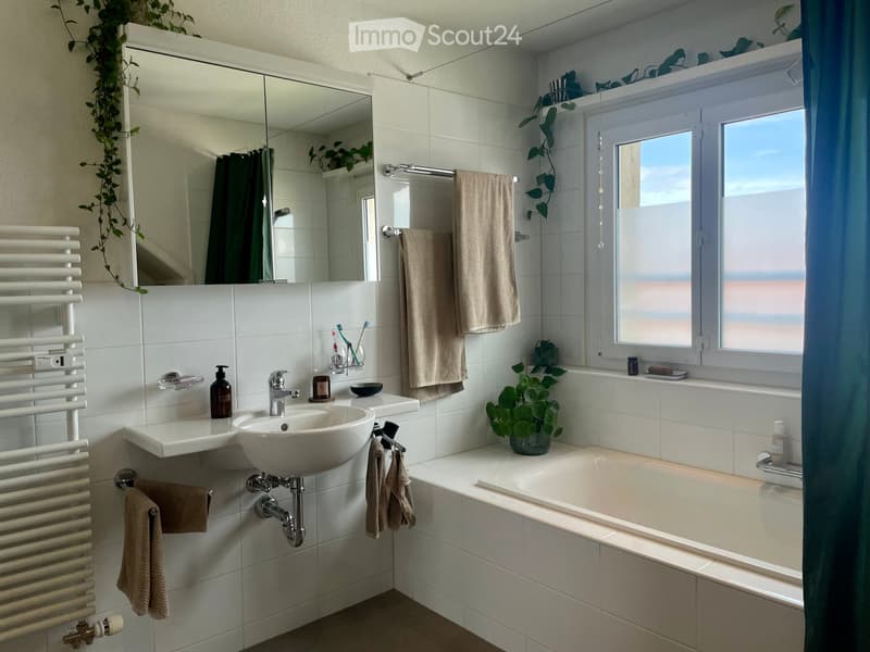 Modern bathroom with white tiles, a large mirror, a sink, a bathtub, and a window with a view. Includes towels, a radiator, and decorative plants.