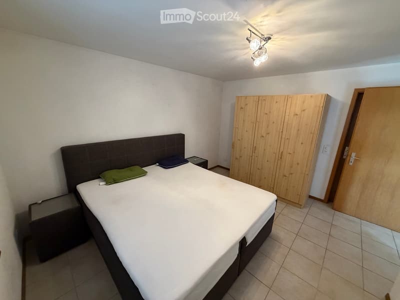 A modern bedroom with a double bed, nightstands, and a large wardrobe. The room features tiled flooring and white walls.