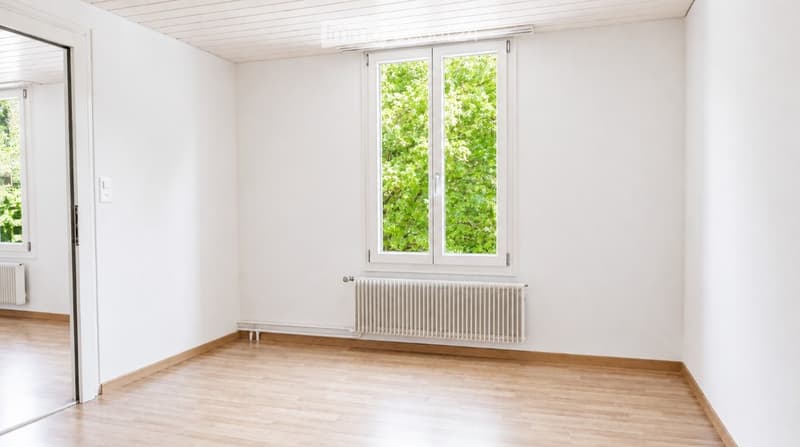 Completely empty room with white walls, wooden floor, and large windows showing greenery outside. No furniture or equipment.
