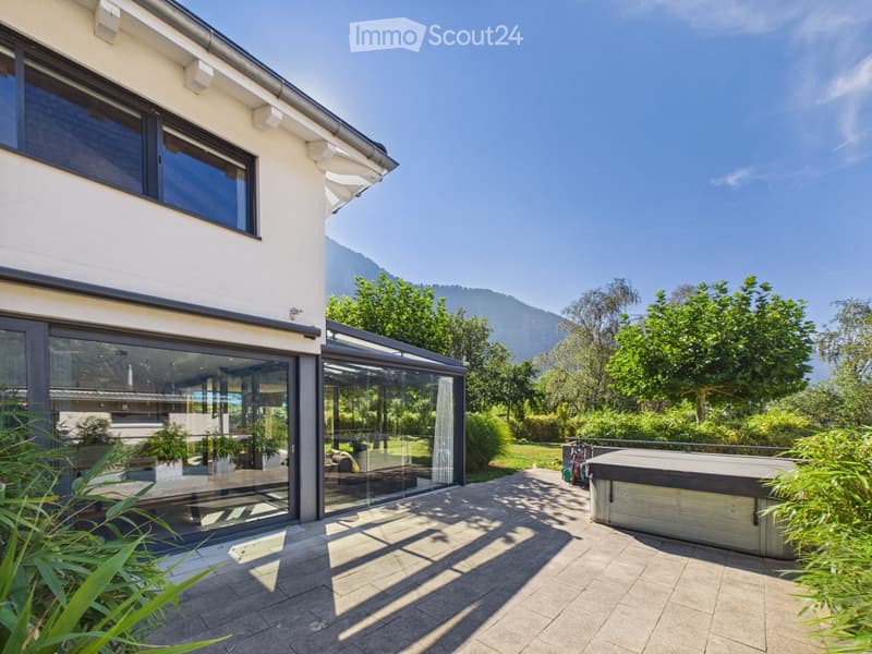 Modern house exterior with large glass sliding doors leading to a patio area, surrounded by lush greenery and a hot tub, with a scenic mountain view in the background.
