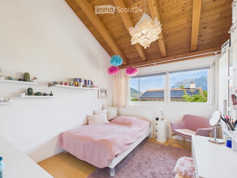 Bright bedroom with wooden ceiling beams, large windows overlooking mountains, white bed with pink bedding, pink sofa, shelves with books and decorative items, and a vanity area with a mirror.