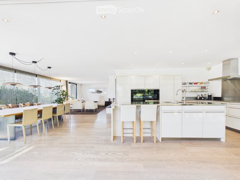 Open-plan kitchen and dining area with white cabinets, a central island, pendant lights, and a dining table with chairs. The space features light wood flooring and large windows with sheer curtains.