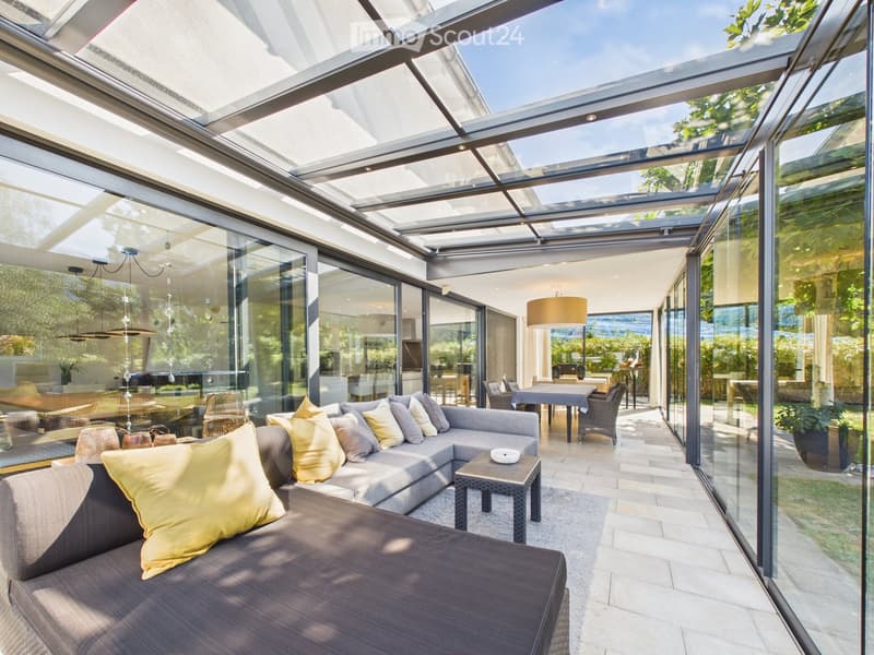Bright, modern living space with large glass walls and a retractable roof, featuring a grey sectional sofa with yellow cushions, a small coffee table, and a dining area with a table and chairs.