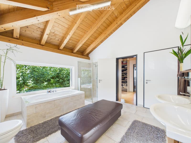 Modern bathroom with a large window offering a view, featuring a bathtub, toilet, double sink, leather stool, and a glass-enclosed shower area.
