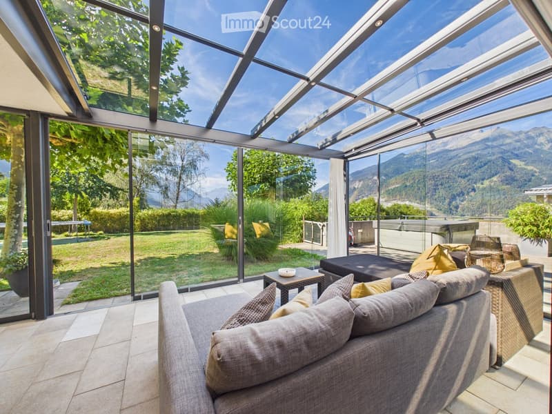 Bright living room with large glass walls and a retractable roof, overlooking a green garden and mountain view. Features a grey sofa, small table, and wicker chairs.