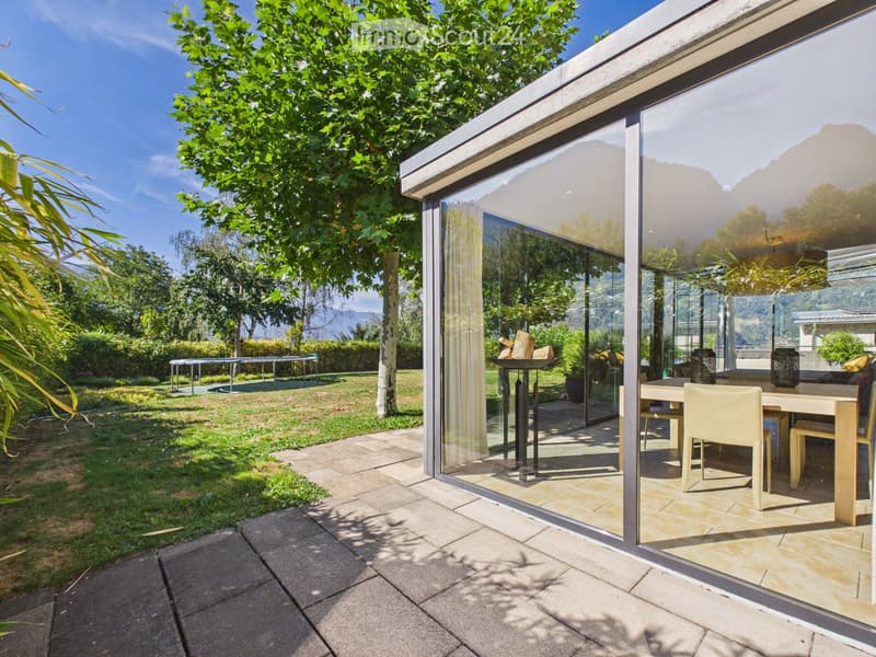 Outdoor patio area with a modern glass extension, featuring a dining table and chairs, surrounded by a well-maintained lawn, trees, and a scenic mountain view.