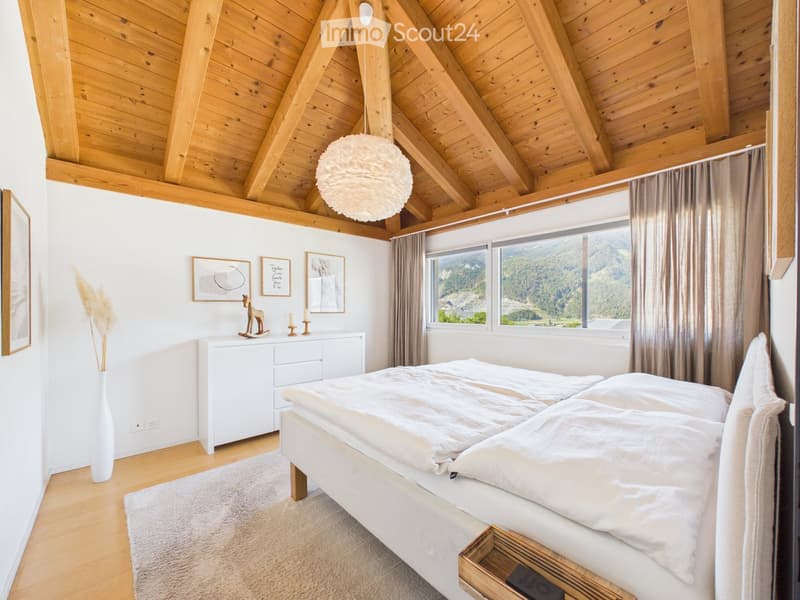 Cozy bedroom with wooden ceiling beams, large windows offering a mountain view, a neatly made bed, a white nightstand, a decorative vase, and framed artwork on the walls.