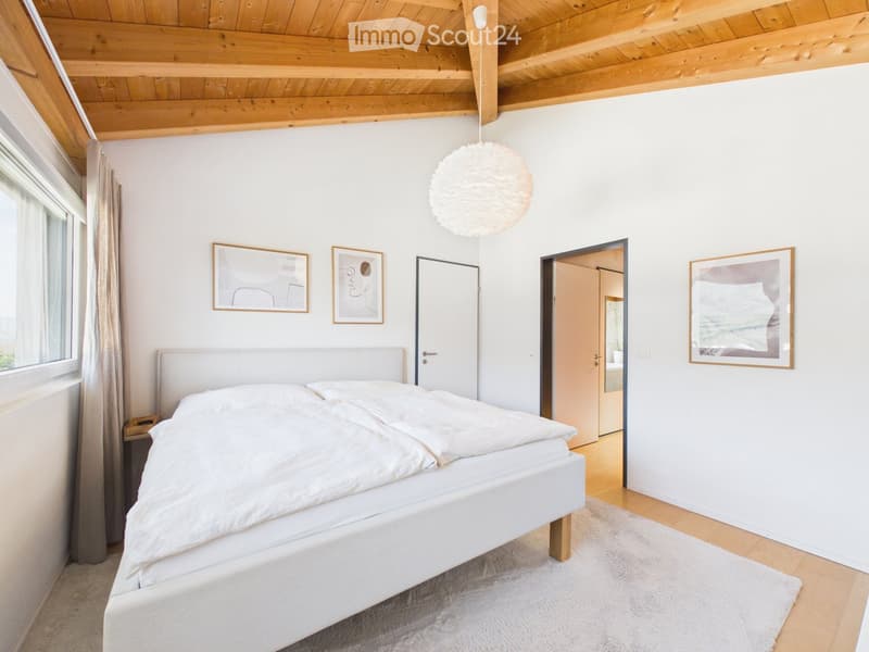 Bright bedroom with a white bed, nightstand, pendant light, and artwork on the walls. The room features wooden flooring and a slanted wooden ceiling.
