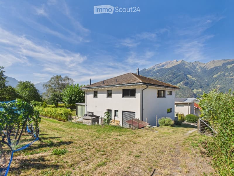 Two-story white house with a brown roof, surrounded by a grassy yard, trees, and mountains in the background.