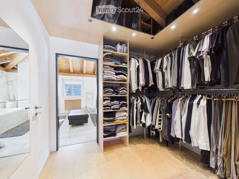 Spacious walk-in closet with wooden flooring, built-in wardrobe, shelves, and an adjacent bathroom with a bathtub and toilet.