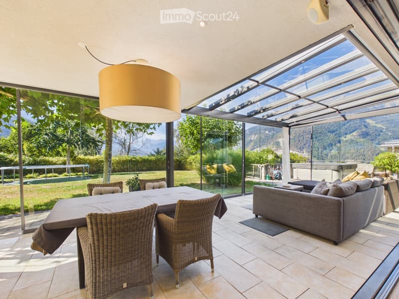 Bright, modern living space with large glass walls offering scenic mountain views, featuring a sofa, dining table, and wicker chairs.
