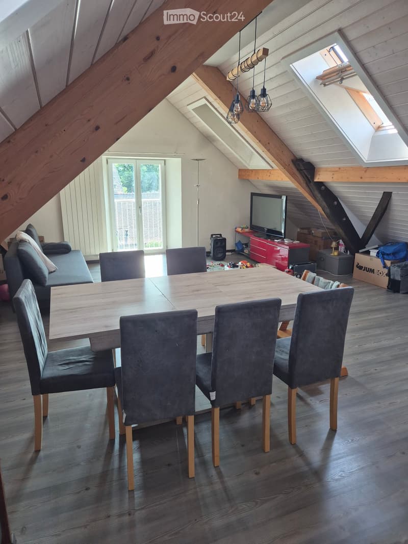 Bright attic room with wooden beams, skylight, dining table with six chairs, sofa, TV, and storage unit.