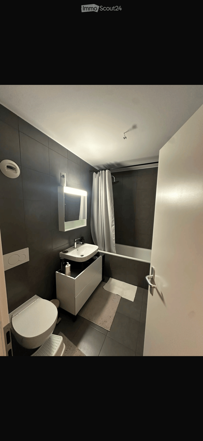 Modern bathroom with dark grey tiles, white fixtures, a toilet, a sink with a mirror, a bathtub with a shower curtain, and a bathroom mat on the floor.