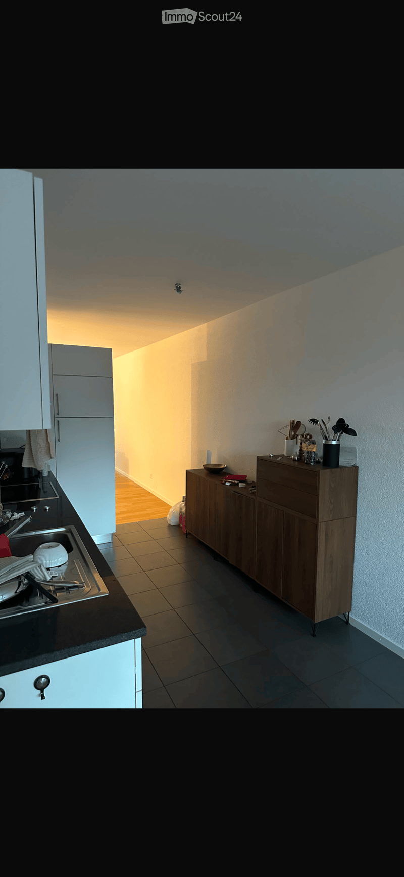 Modern kitchen with white cabinets, black countertop, and stainless steel sink. Wooden floor and tiled walls visible.