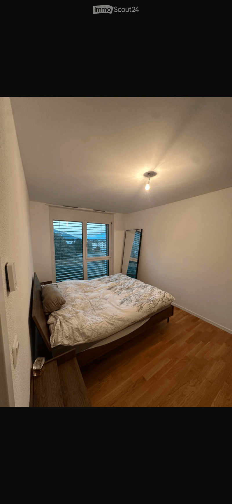 Bedroom with wooden flooring, a bed with a patterned duvet, a mirror, and a window with blinds.
