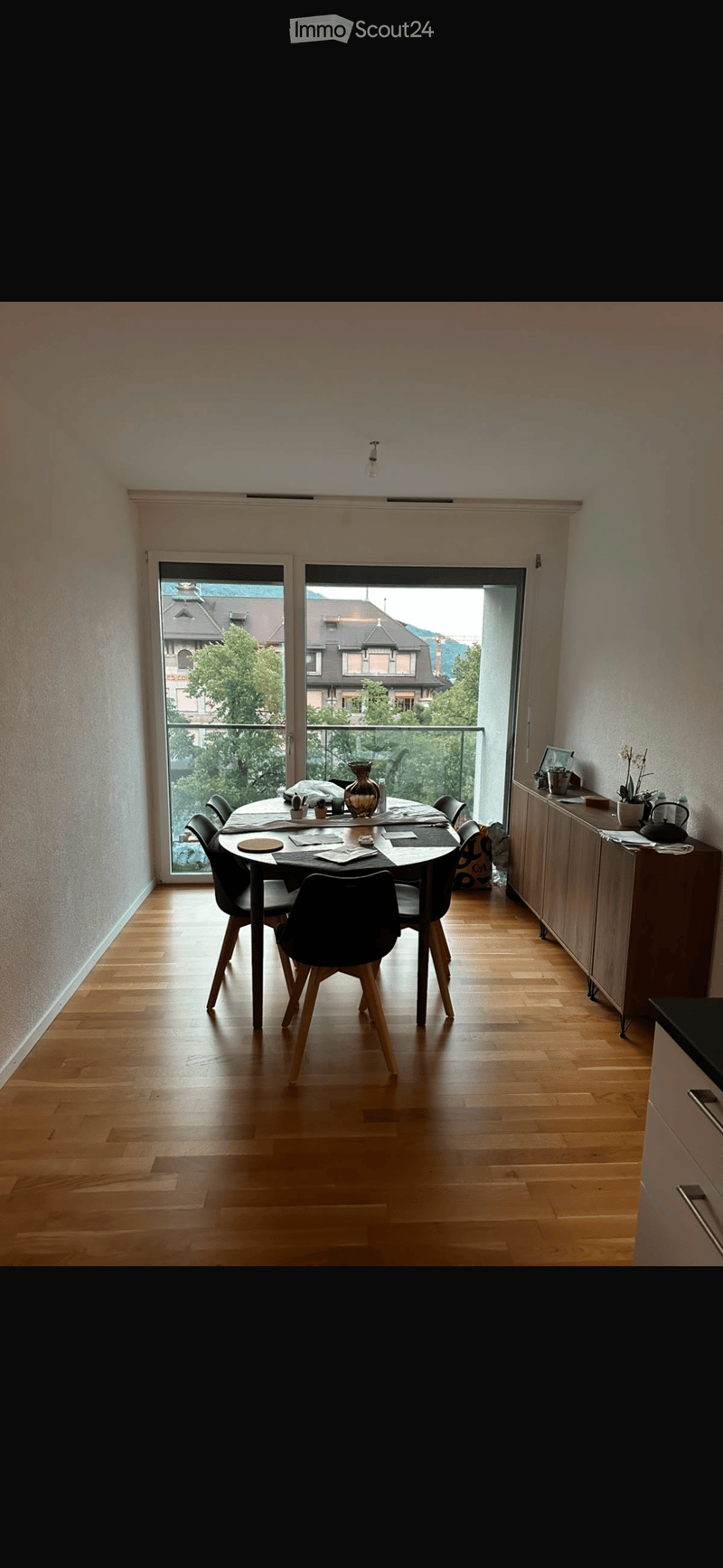 Dining room with wooden floor, large window, dining table with chairs, vase, and a small cabinet with a teapot and other items.