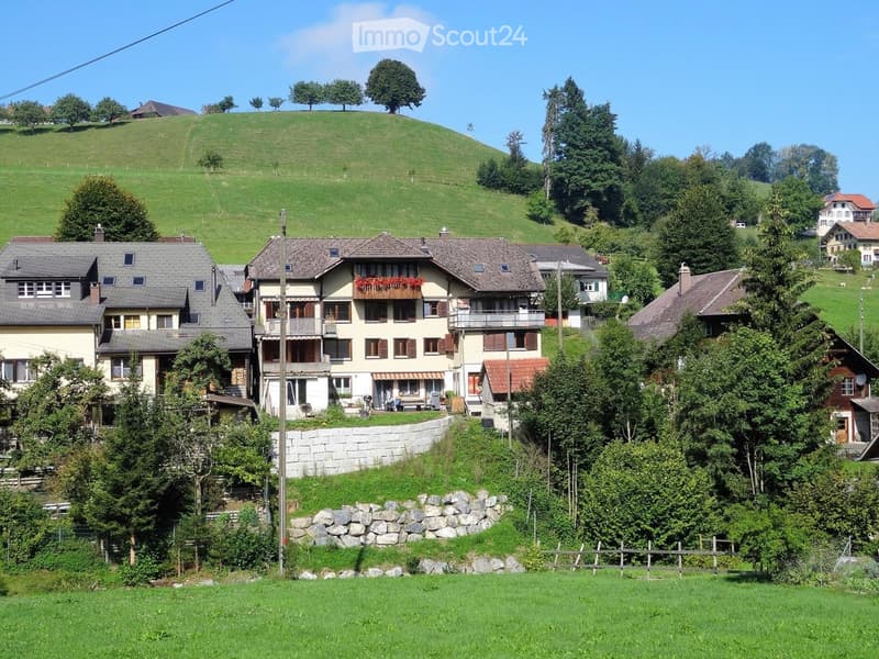 A cluster of traditional houses with pitched roofs and balconies, surrounded by lush green hills and trees.