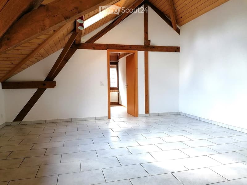 Empty attic space with exposed wooden beams, white walls, tiled floor, and a doorway leading to another room.