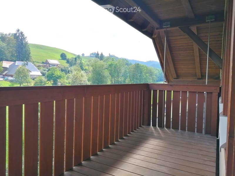 Wooden balcony with a railing, overlooking a green, hilly landscape with trees and houses.