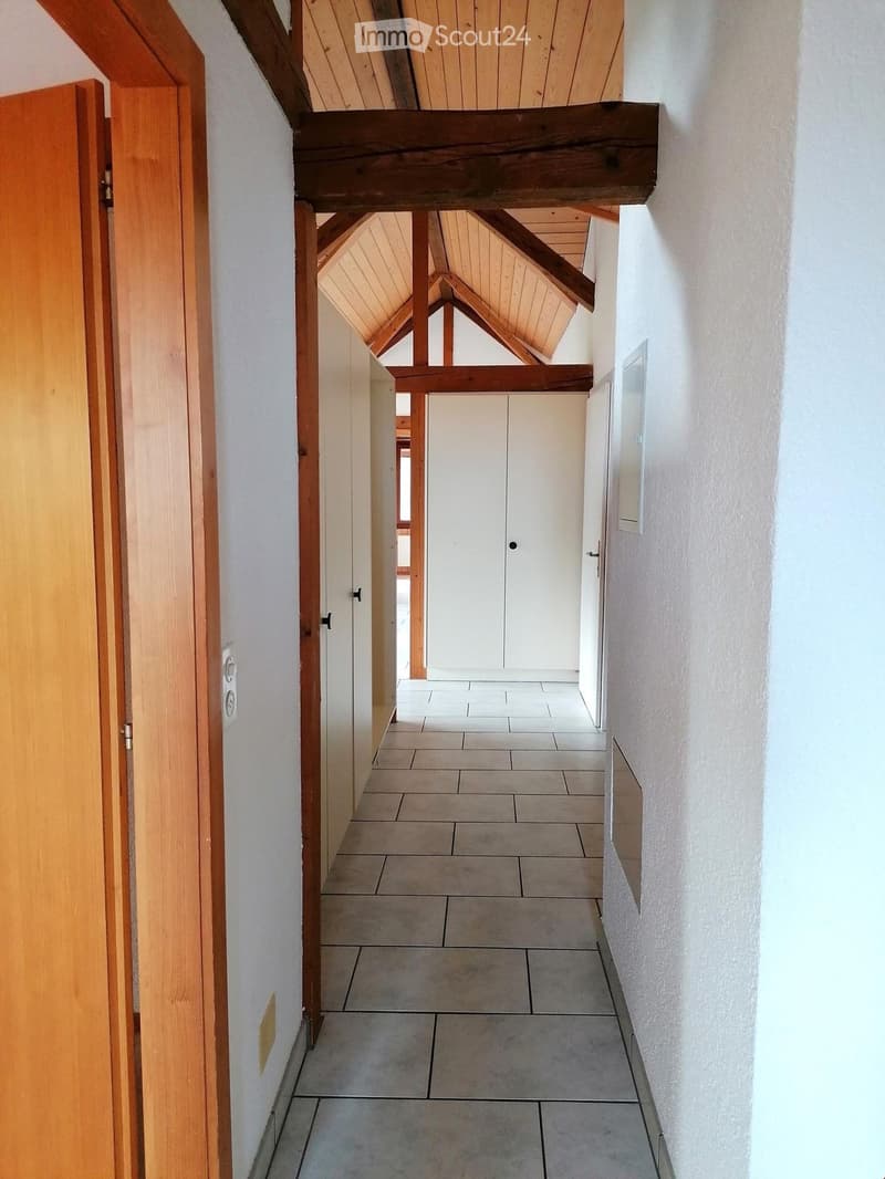 A narrow hallway with exposed wooden beams and a staircase leading upwards, featuring tiled flooring and white walls.