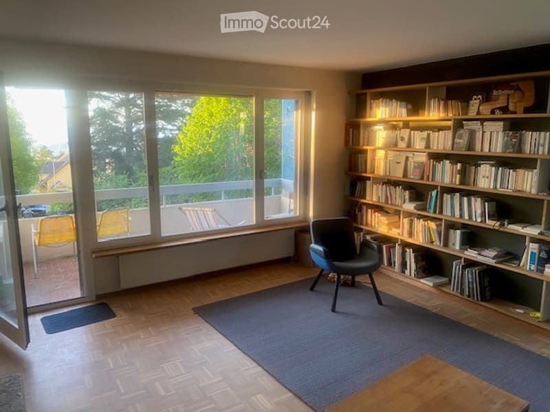 Bright living room with large windows and a balcony, featuring a wooden floor, a blue armchair, a bookshelf filled with books, and a small wooden table with a rug.