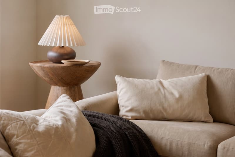 A modern living room with a beige sofa, decorative pillows, a wooden side table, and a table lamp with a white shade.
