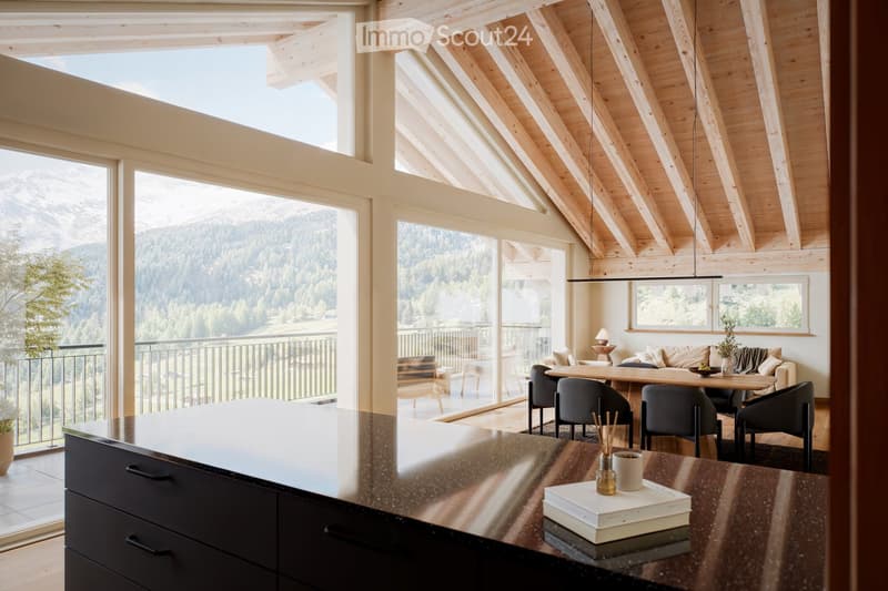 Open-plan living and dining area with exposed wooden beams, large windows offering a mountain view, a kitchen island, a dining table with chairs, a sofa, and decorative items on the counter.