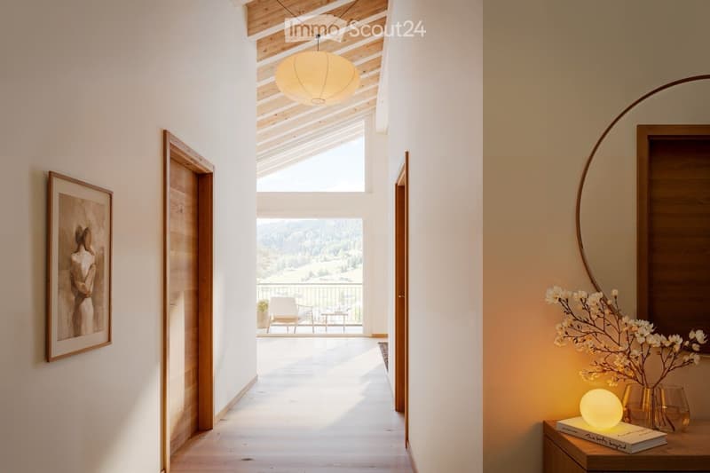 Bright hallway with wooden flooring, white walls, and a large skylight. Features a round mirror, a small table with a lamp and decorative items, and a framed artwork on the wall. The hallway leads to a balcony area with outdoor seating.